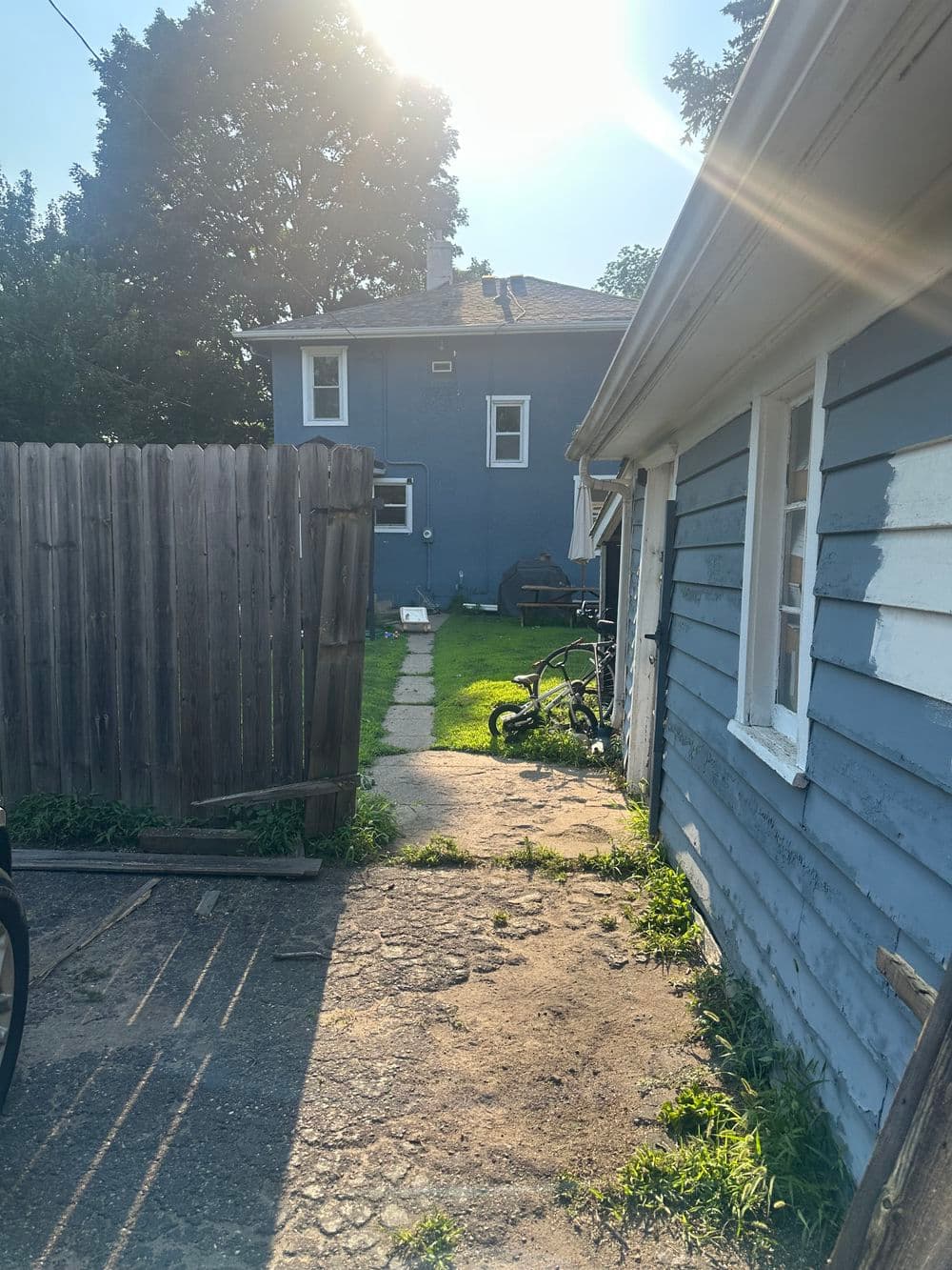 Narrow pathway beside blue house, leading to backyard with sunlight and green grass.