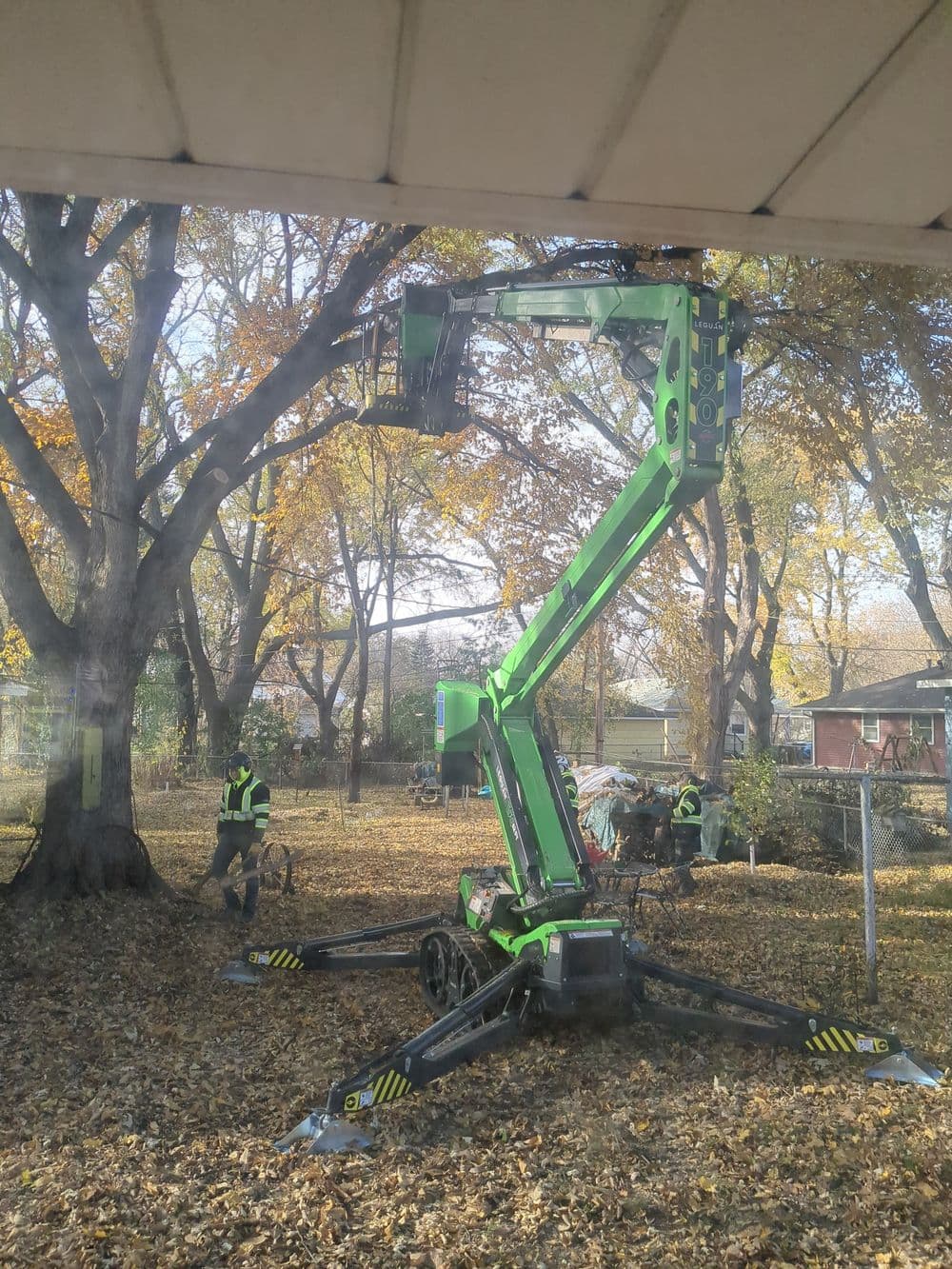 A green cherry picker lifting workers for tree maintenance in a leaf-covered yard.