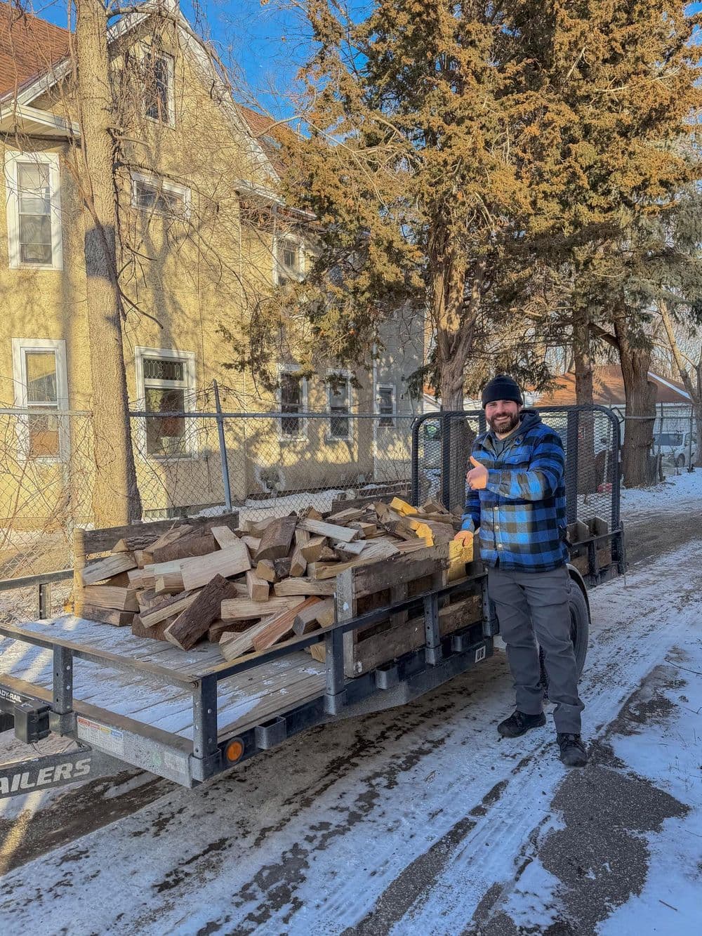 Person in a blue checkered shirt stands beside a trailer loaded with firewood in winter.