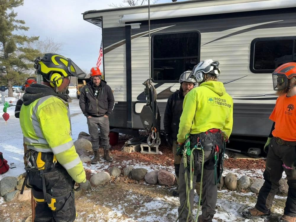 Tree service professionals in safety gear discussing equipment near a camper in winter setting.