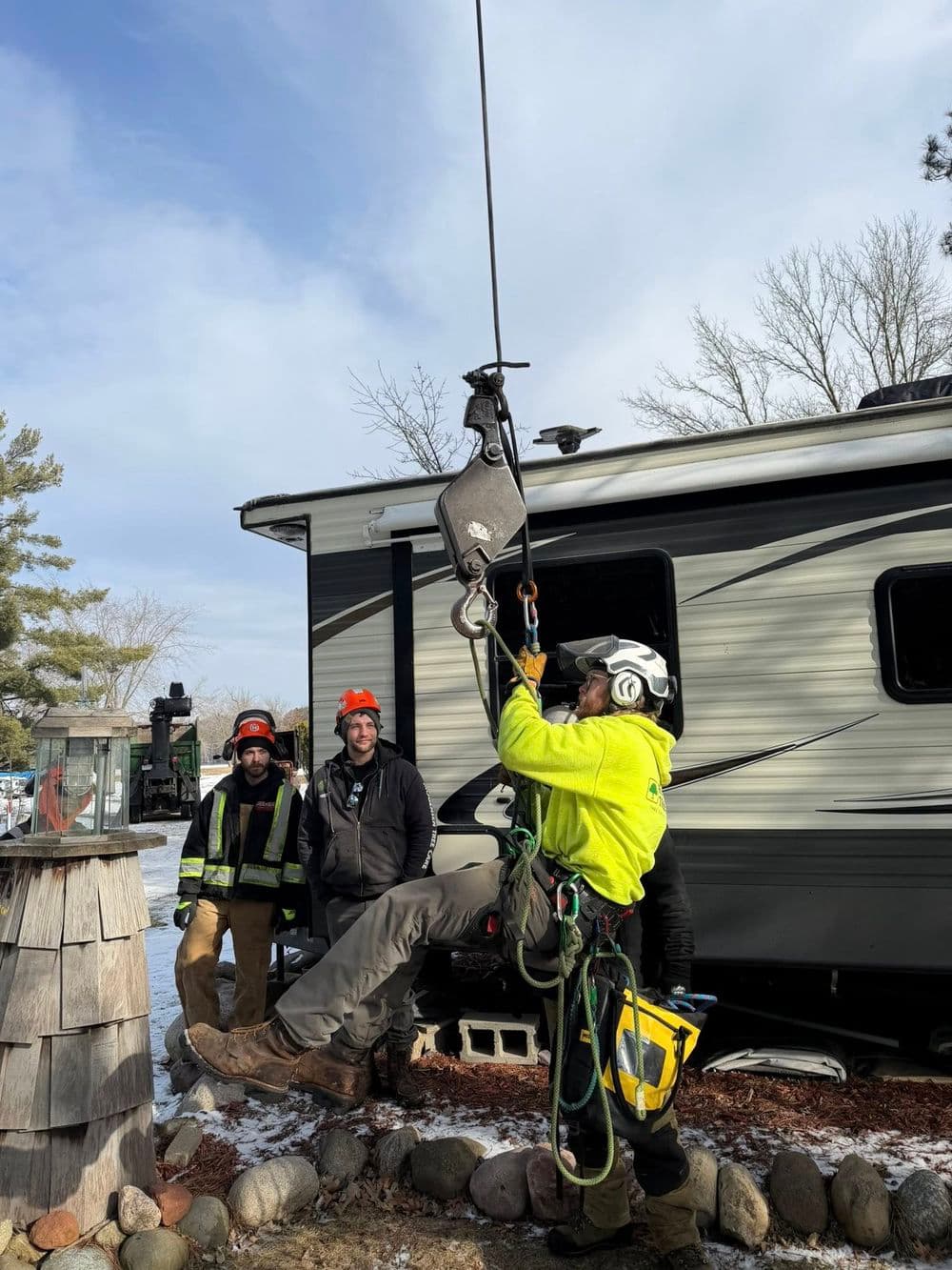 Worker using a pulley system to lift equipment near an RV, with three observers in safety gear.